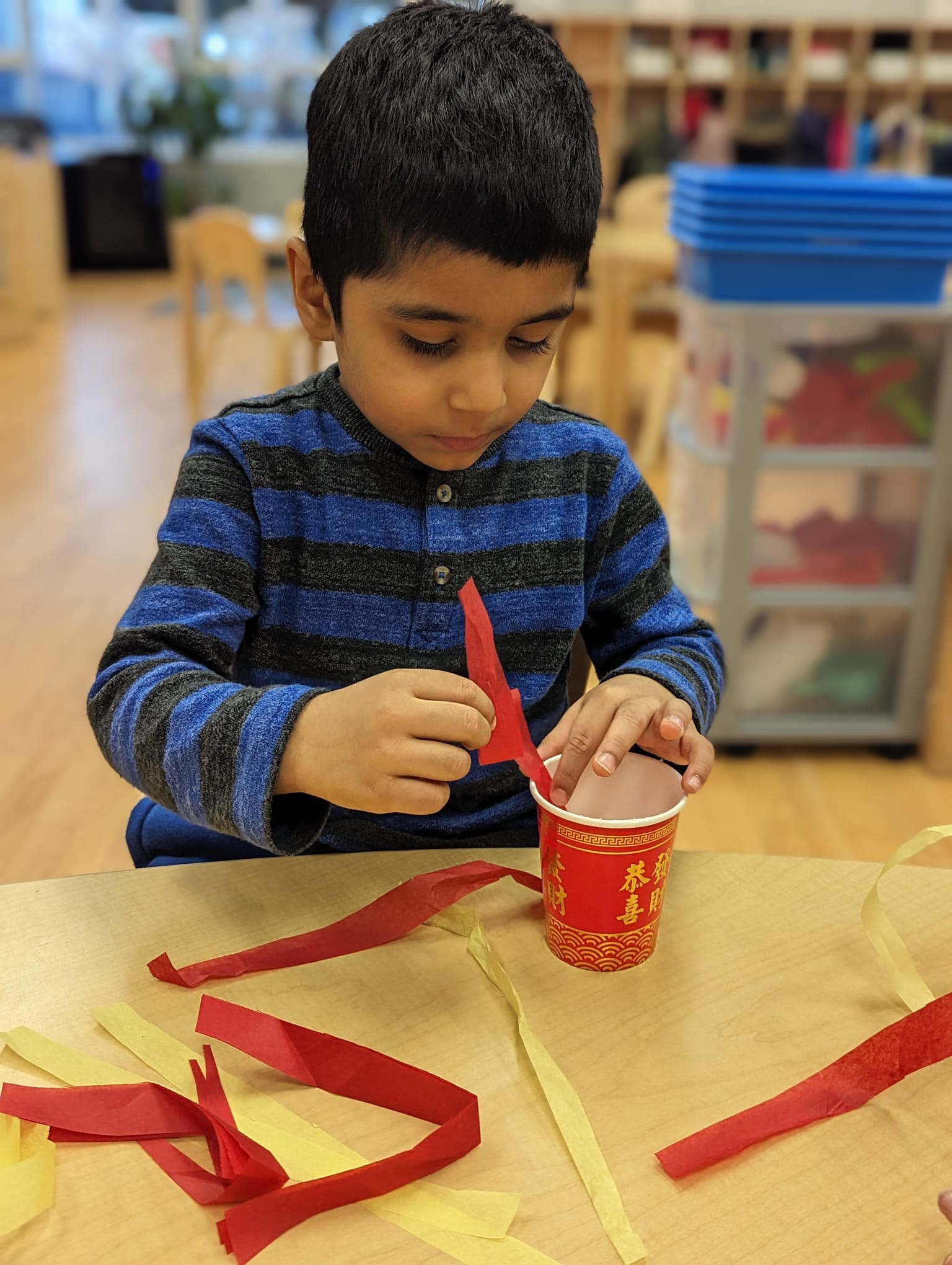 A young boy in a PLASP Early Learning and Child Care Centre sits at a table in a PLASP Early Learning and Child Care Centre and makes a craft with yellow and red ribbon for Lunar New Year